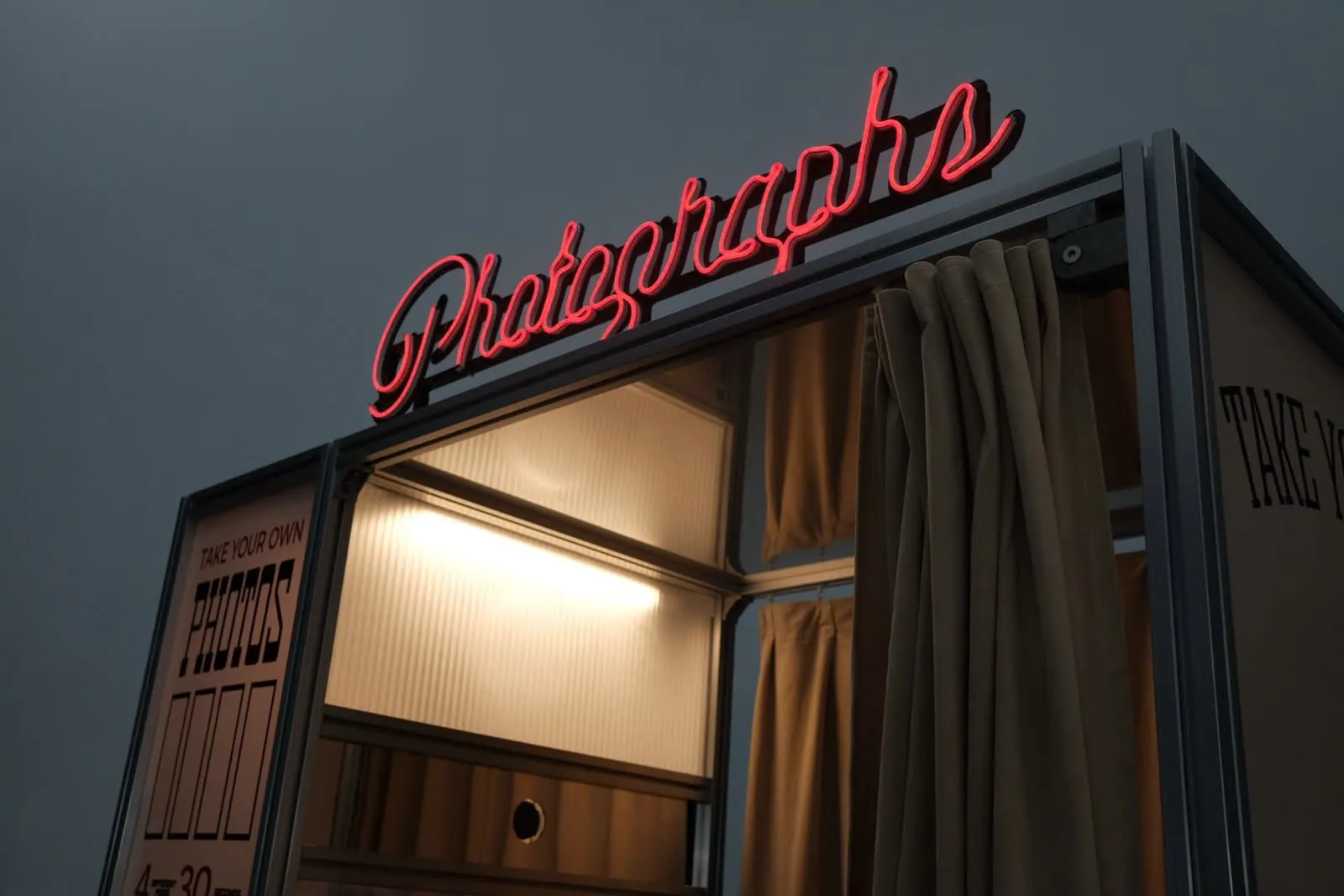 Close-up of the glowing red Photographs neon sign above the vintage photo booth entrance at dusk